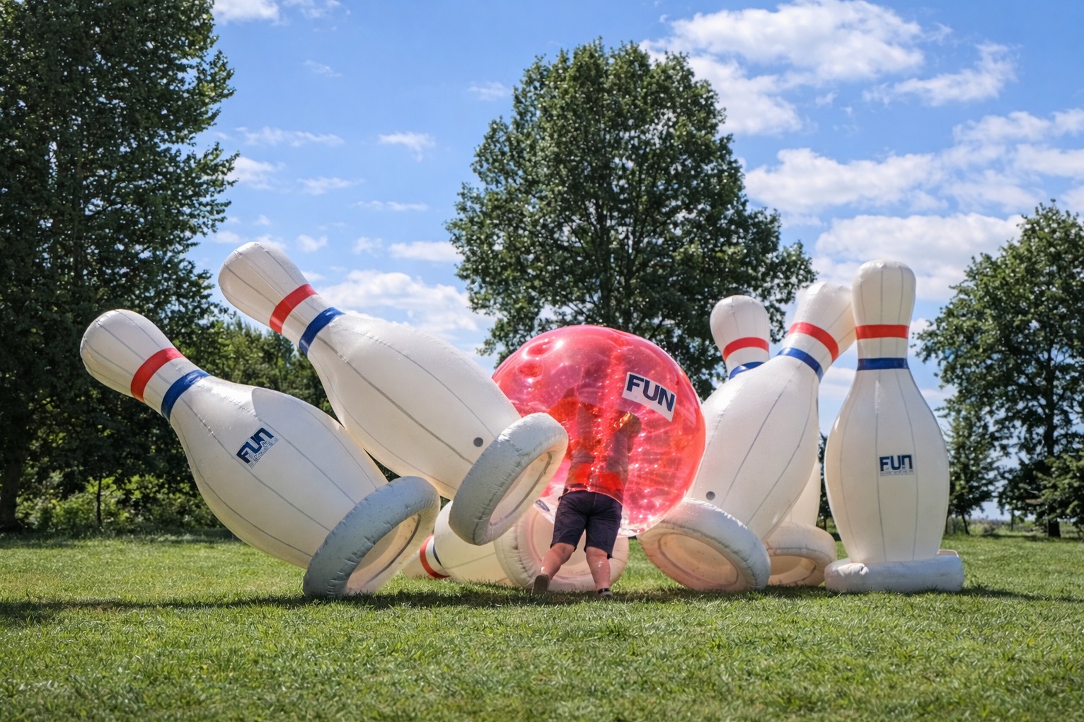 Dmuchane kręgle i kula z osobą w środku na trawie, w tle drzewa i błękitne niebo. Atrakcja eventowa z logo 'FUN'.