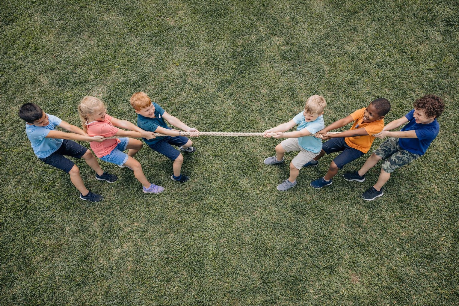 Zabawa w przeciąganie liny na zielonej trawie. Grupa dzieci w różnym wieku i ubraniach rywalizuje w grze zespołowej. Widok z góry, dynamiczne ujęcie.