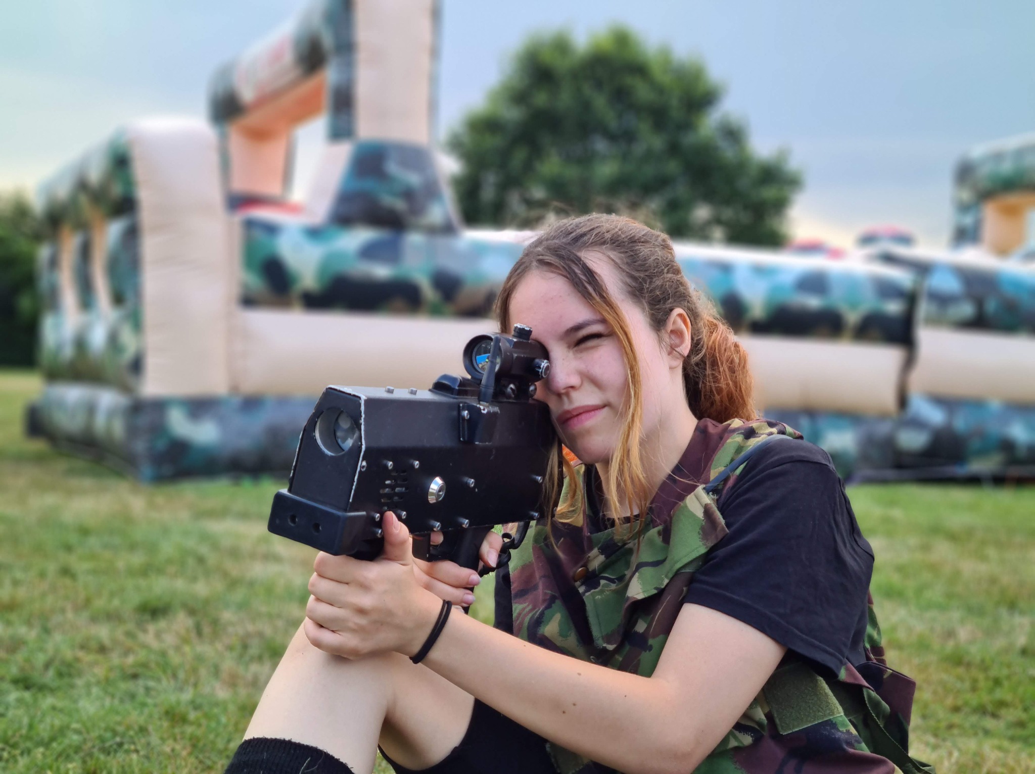 Uśmiechnięta dziewczyna celuje z laserowego pistoletu na tle dmuchanego labiryntu w kamuflażu, siedząc na trawie. Gra laser tag na imprezie plenerowej.
