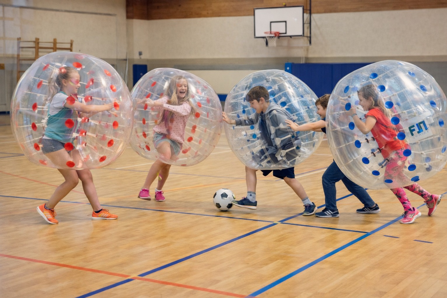 Dzieci w przezroczystych kulach grają w piłkę nożną na hali sportowej z drewnianą podłogą i koszem do koszykówki w tle. Aktywna zabawa.