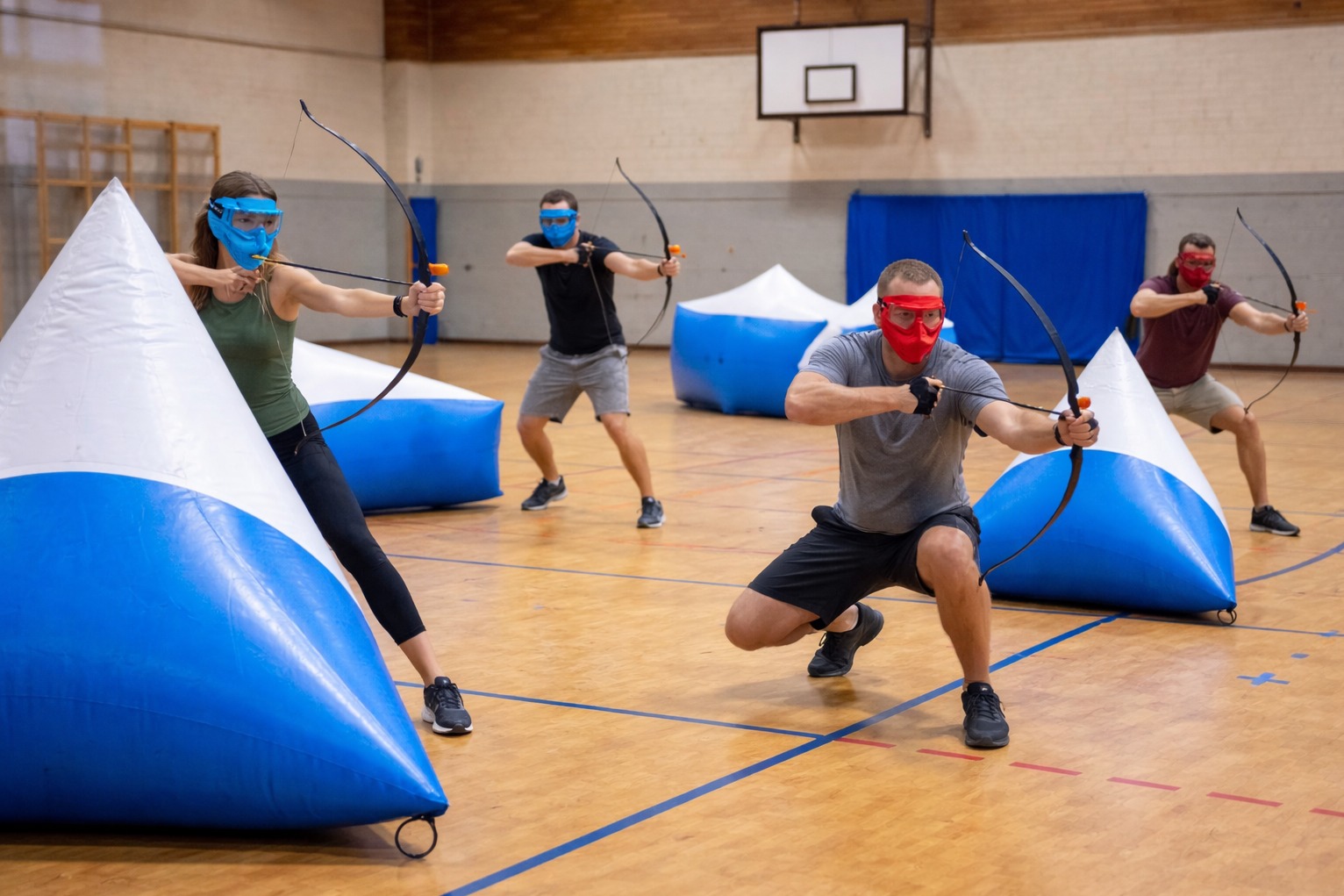 Gra w Archery Tag: grupa osób w maskach i z łukami celuje do dmuchanych przeszkód na sali gimnastycznej. Dynamiczne ujęcie z perspektywy uczestnika.