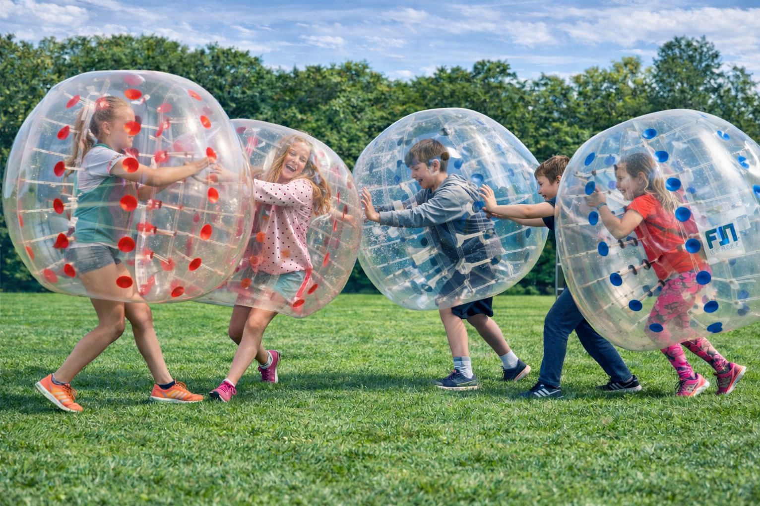 Grupa dzieci w przezroczystych kulach bumper ball na zielonej trawie, zabawa na świeżym powietrzu, dynamiczne ujęcie z perspektywy poziomej.