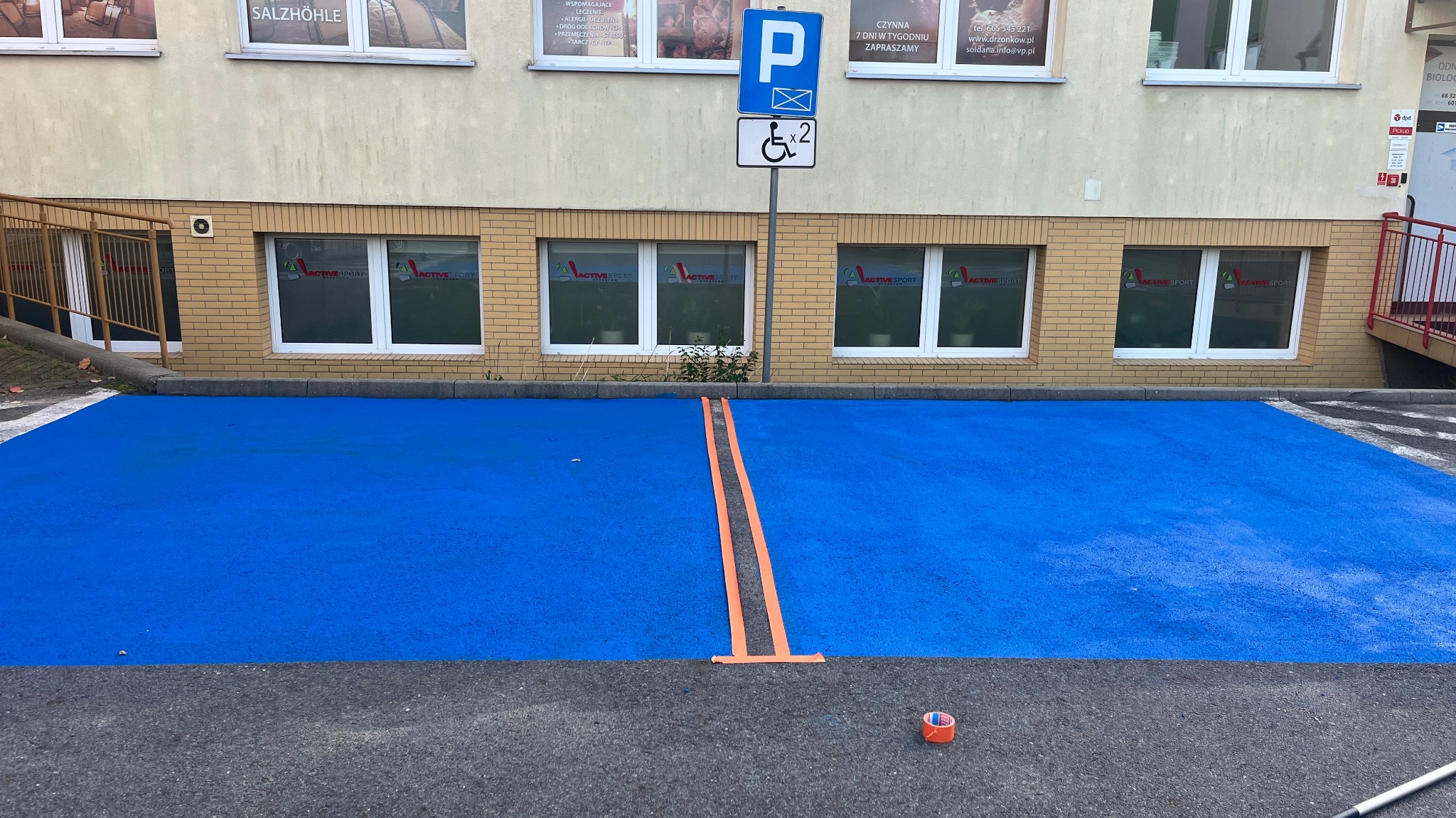 Niebieskie miejsce parkingowe dla niepełnosprawnych, wyznaczone pomarańczową taśmą, przed budynkiem z oknami i znakiem P. Widoczna rolka taśmy.