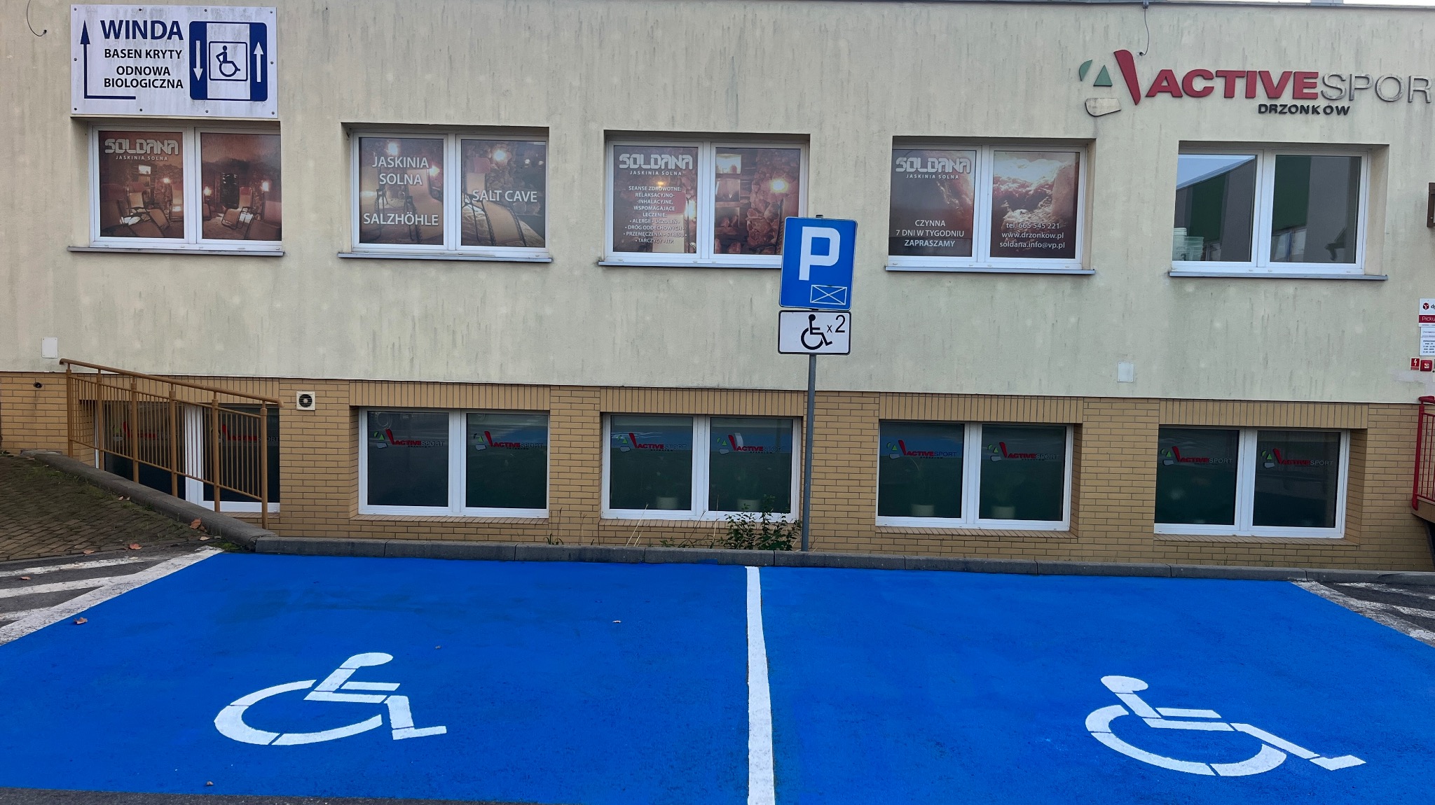 Budynek Active Sport z oknami reklamującymi Jaskinię Solną Soldana i miejscami parkingowymi dla niepełnosprawnych. Widoczny szyld i reklamy w oknach.