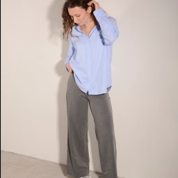 Anna Jachimczak - Kobieta z kręconymi włosami w błękitnej koszuli i szarych spodniach pozuje na jasnym tle. Ręka w kieszeni, wzrok skierowany w dół. Casualowy styl.