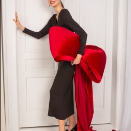 Anna Jachimczak - Uśmiechnięta kobieta w czarnej sukience pozuje z dużym, czerwonym, aksamitnym rekwizytem przypominającym kokardę na tle białych drzwi.