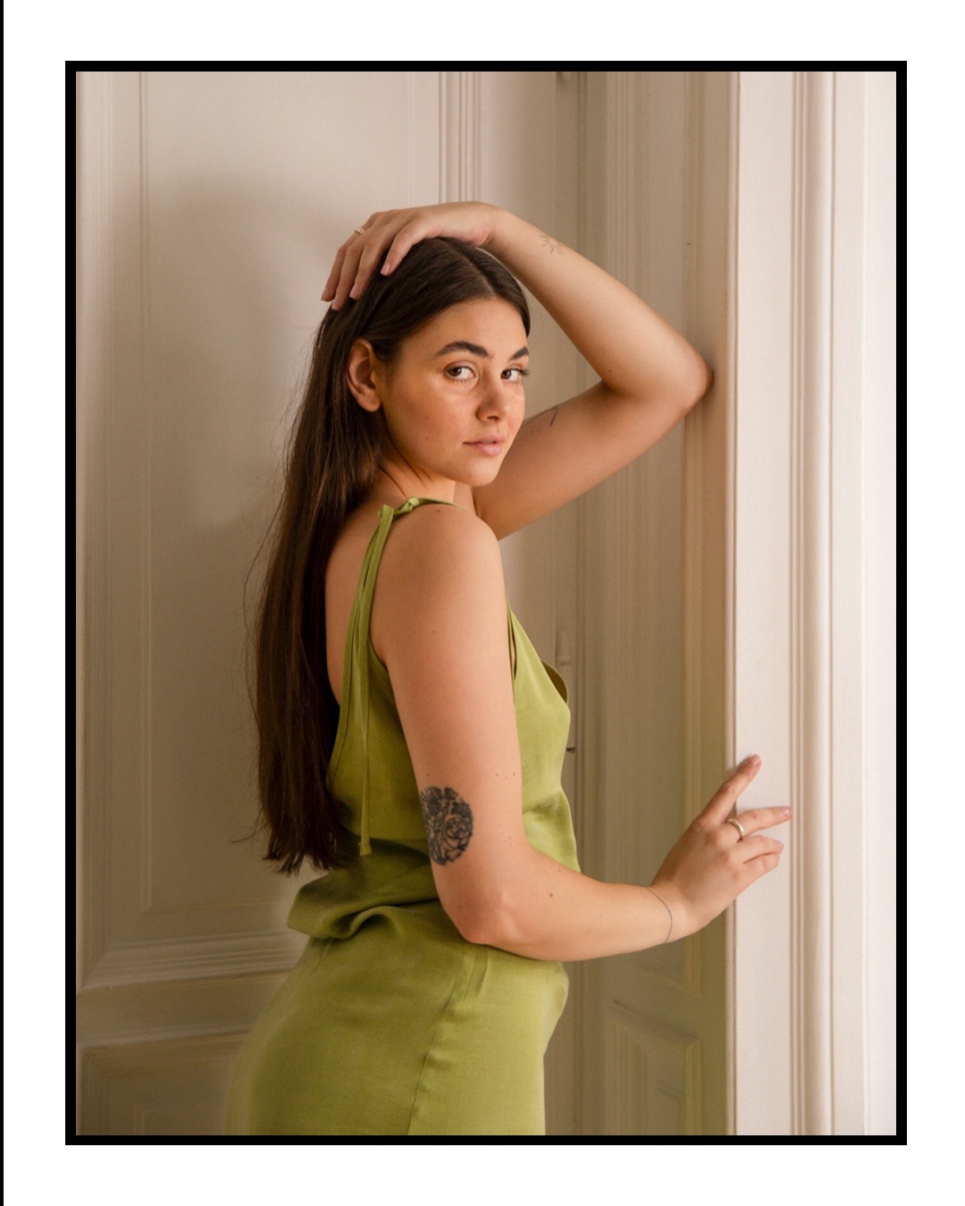Portret kobiety w zielonej sukience, opierającej się o białe drzwi. Widoczny tatuaż na ramieniu i pierścionki na palcach. Naturalne światło podkreśla rysy twarzy.