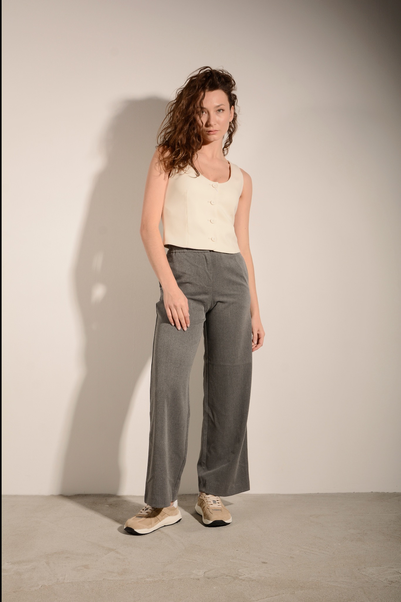 Portret kobiety w beżowej kamizelce i szarych spodniach na tle jasnej ściany. Ujęcie w pełnej sylwetce, z delikatnym oświetleniem i naturalną mimiką.