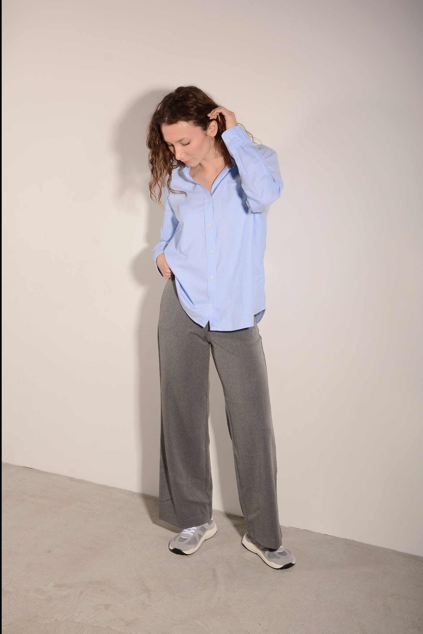 Kobieta z kręconymi włosami w błękitnej koszuli i szarych spodniach pozuje na jasnym tle. Ręka w kieszeni, wzrok skierowany w dół. Casualowy styl.