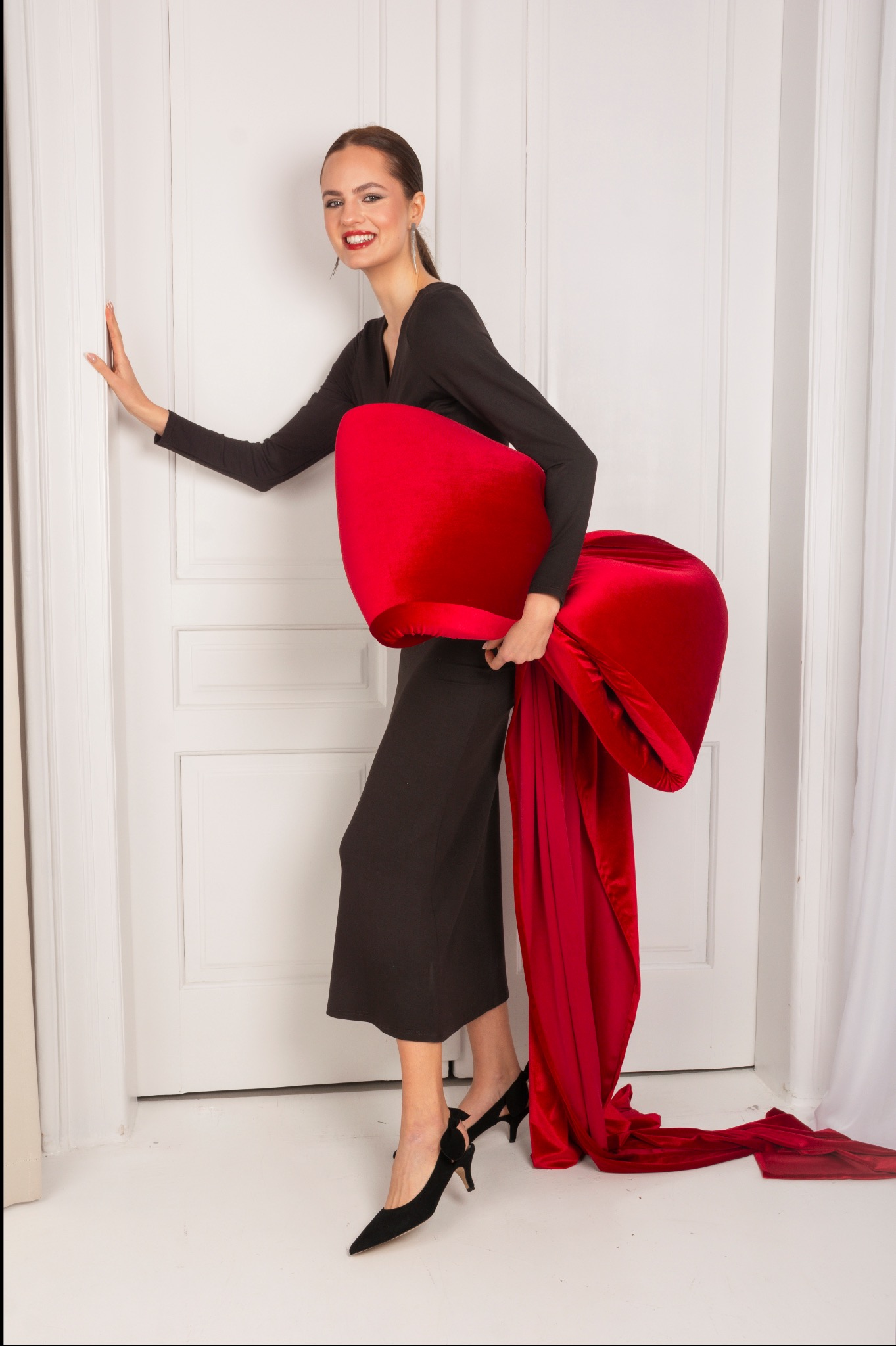 Uśmiechnięta kobieta w czarnej sukience pozuje z dużym, czerwonym, aksamitnym rekwizytem przypominającym kokardę na tle białych drzwi.