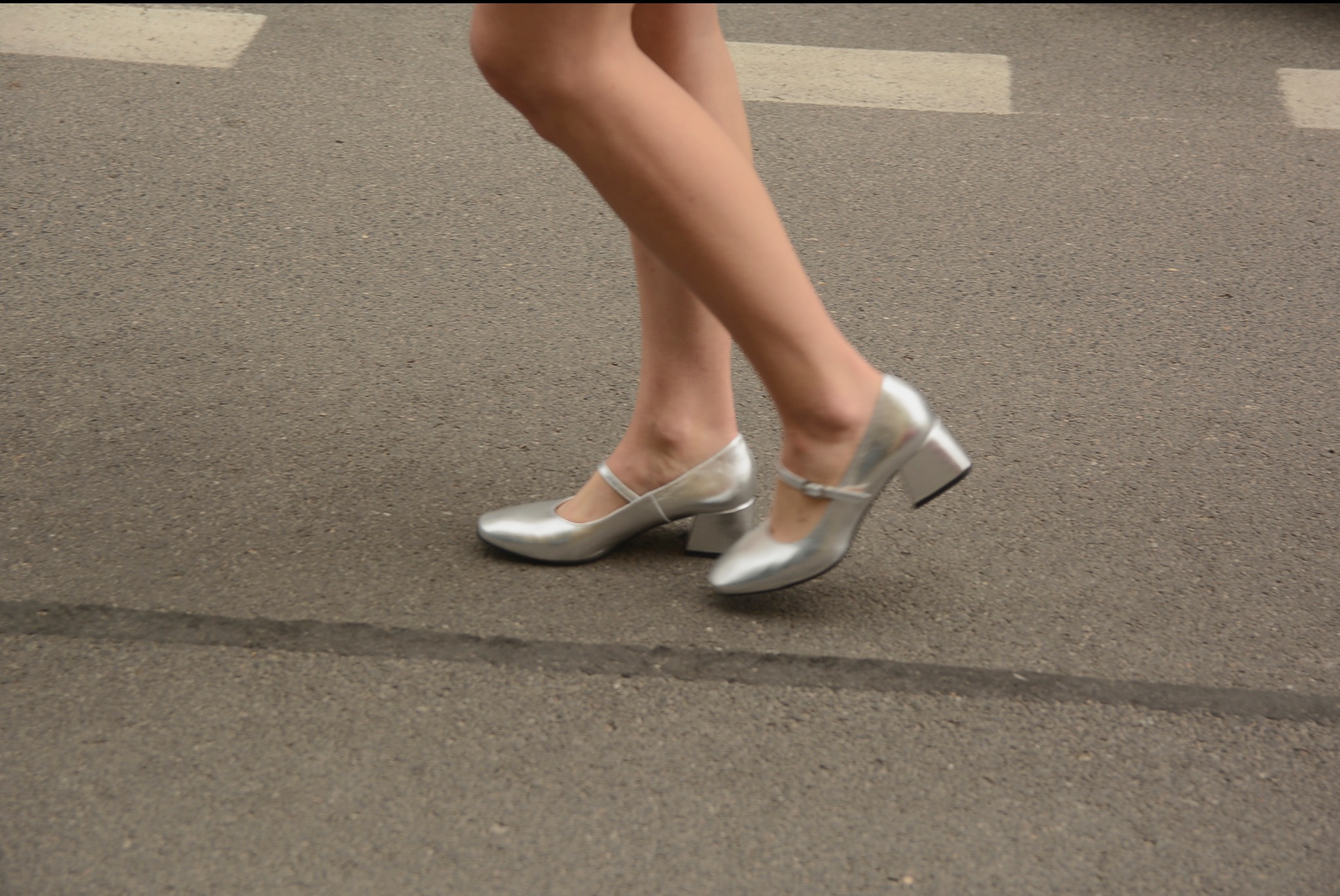 Nogi w srebrnych, błyszczących butach na niskim obcasie, na tle szarego asfaltu z widocznymi liniami. Ujęcie od dołu, skupione na obuwiu i dolnej części nóg.