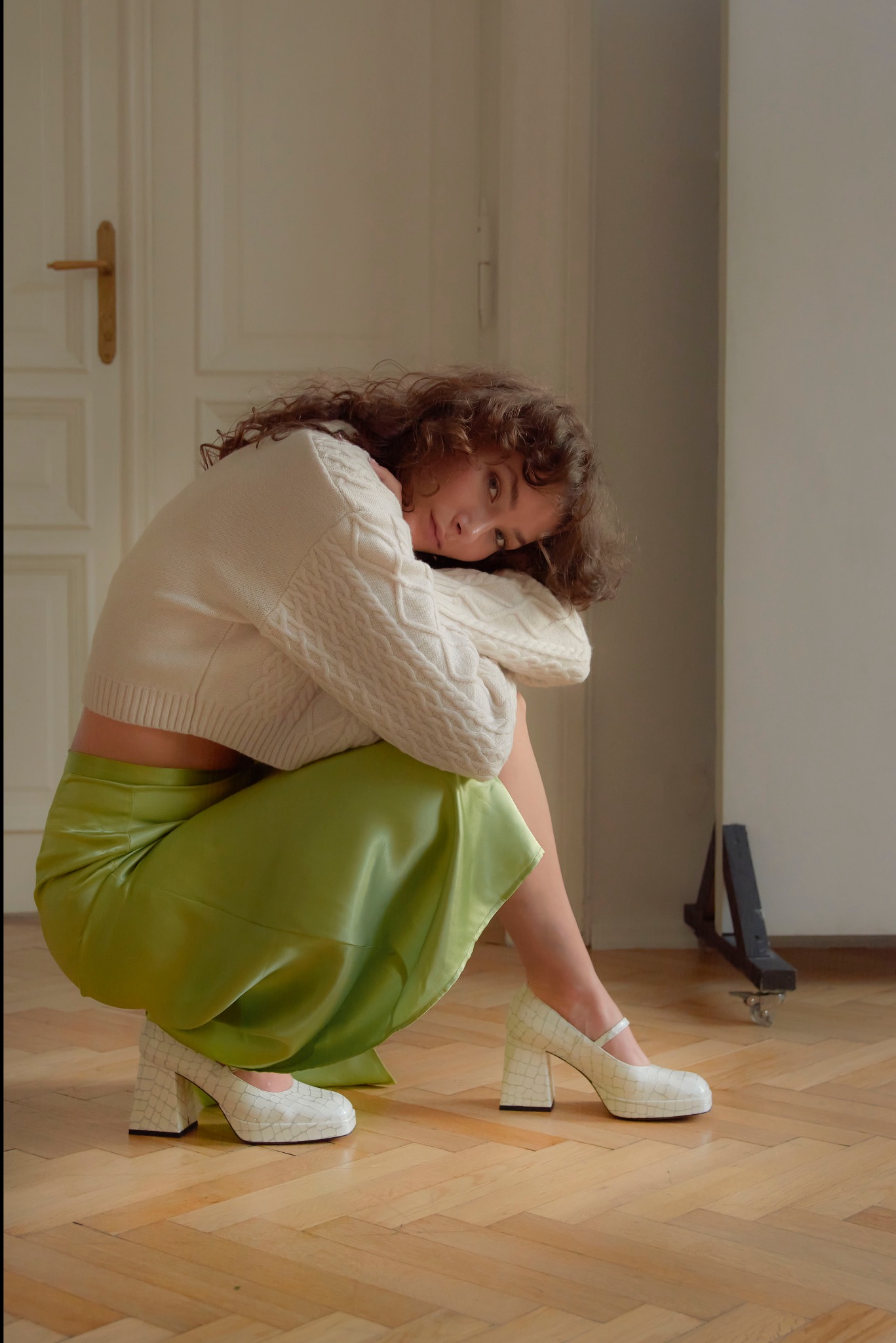 Kobieta w białym swetrze i zielonej spódnicy pozuje w kucki, opierając głowę na kolanach. Uwagę przyciągają białe buty na platformie i obcasie. W tle widoczne drzwi i drewniana podłoga.