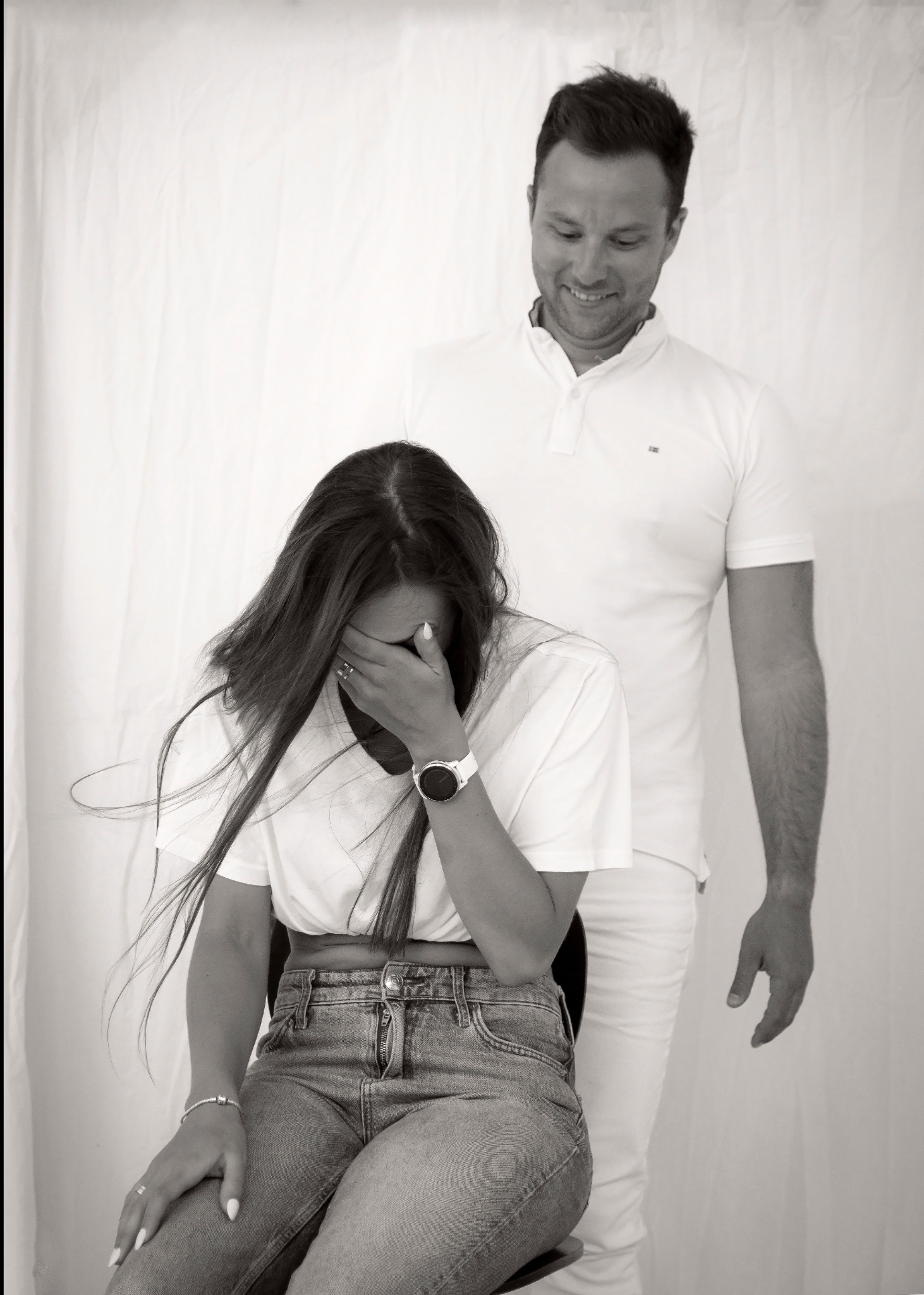 Czarno-białe zdjęcie pary: kobieta siedzi zakrywając twarz, mężczyzna stoi za nią z uśmiechem. Ubrani w jasne ubrania na tle białej tkaniny.