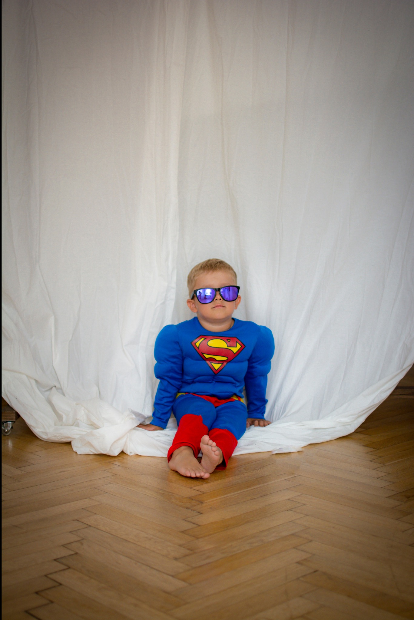 Chłopiec w kostiumie Supermana i okularach przeciwsłonecznych siedzi na podłodze z parkietu na tle białej tkaniny. Ujęcie z dołu, podkreślające dziecięcą perspektywę.