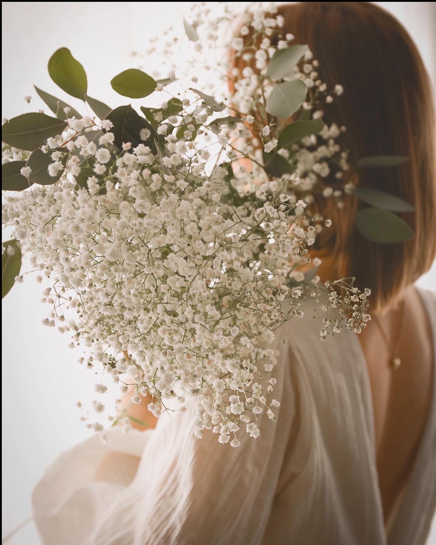 Kobieta z bukietem gipsówki i eukaliptusa, widok z tyłu, delikatne światło, subtelna kolorystyka, minimalistyczny styl, naszyjnik z zawieszką.