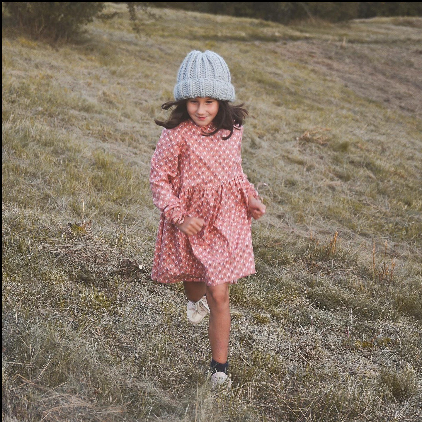 Dziewczynka w różowej sukience i szarej czapce biegnie po trawiastym wzgórzu. Uśmiechnięta, w ruchu, z rozwianymi włosami. Naturalne światło, plener.
