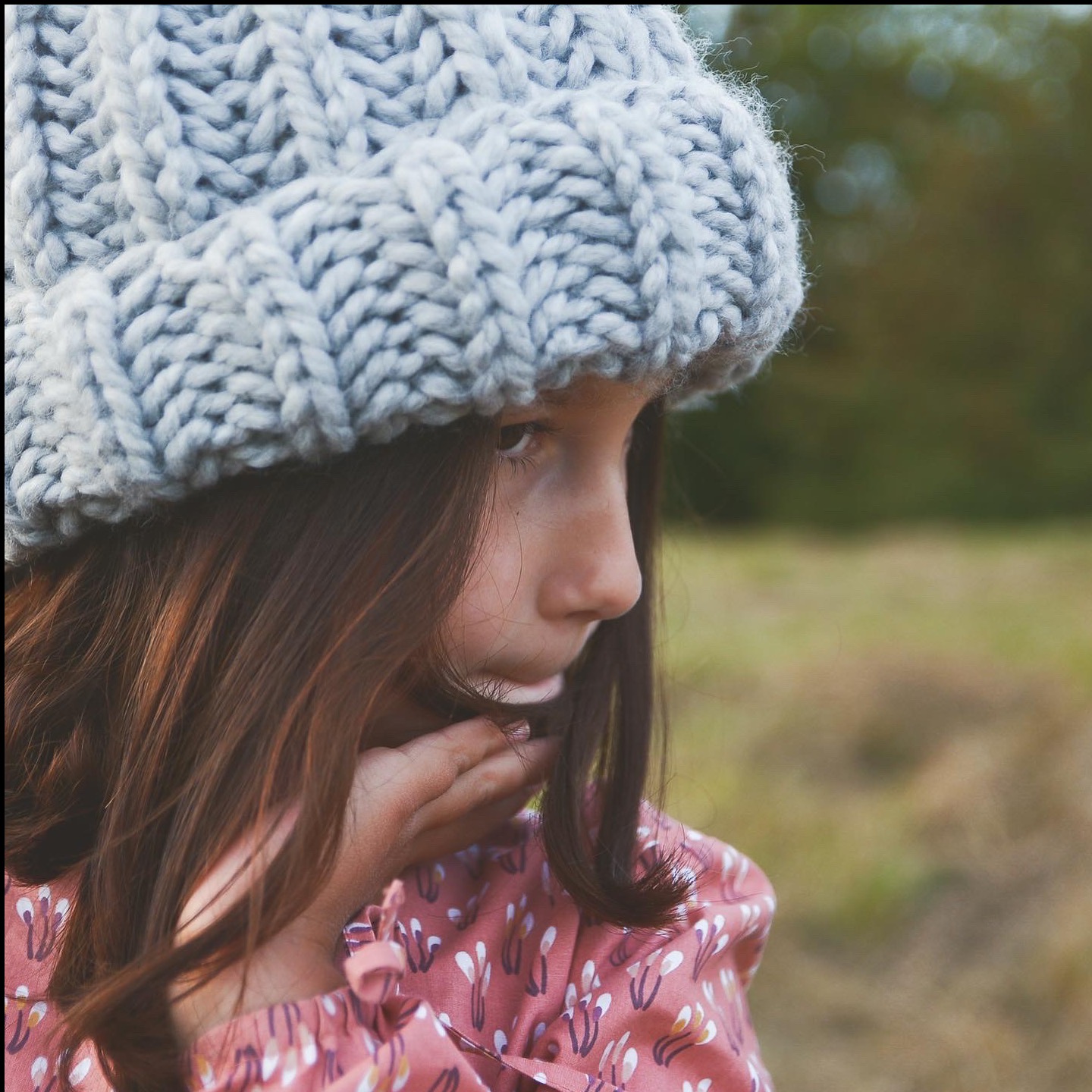 Profil dziewczynki w wełnianej czapce, zamyślona poza na tle zieleni. Delikatne światło podkreśla detale dzierganej czapki i wzór na bluzce.