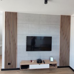 AS Serwis - Nowoczesny salon z szarą ścianą z płytek, telewizorem, drewnianymi lamelami po bokach i szafką RTV. Podłoga z paneli imitujących drewno. Minimalistyczne oświetlenie.