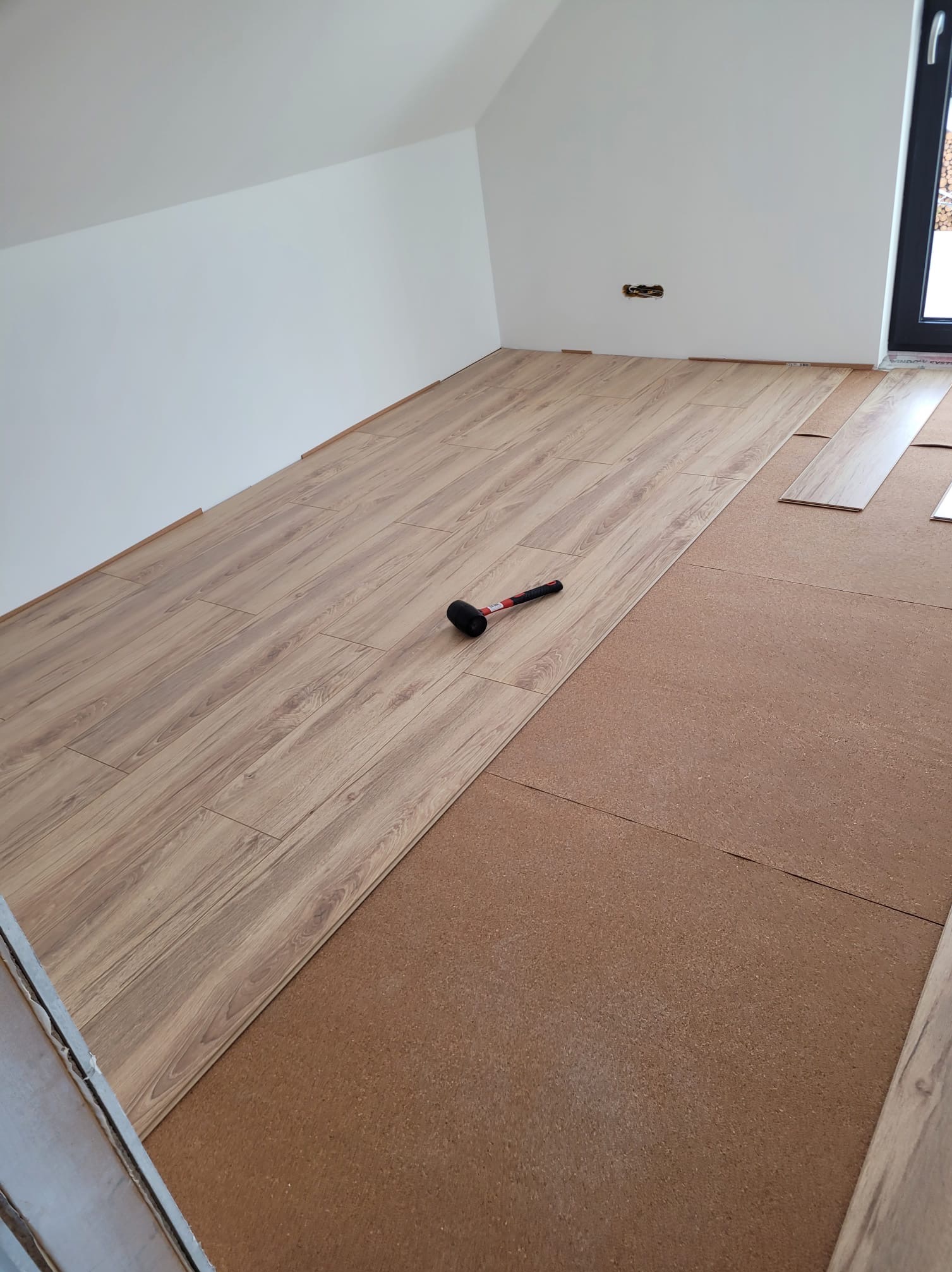 Układanie paneli podłogowych w jasnym pomieszczeniu na podkładzie korkowym. Widoczny gumowy młotek i fragment okna. Częściowo ułożona podłoga.