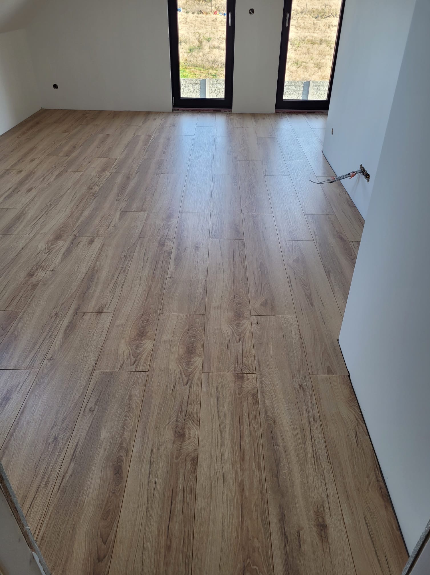 Ukończona podłoga z paneli drewnopodobnych w jasnym wnętrzu z oknami. Widoczne gniazdka elektryczne na ścianie. Perspektywa od wejścia do pokoju.