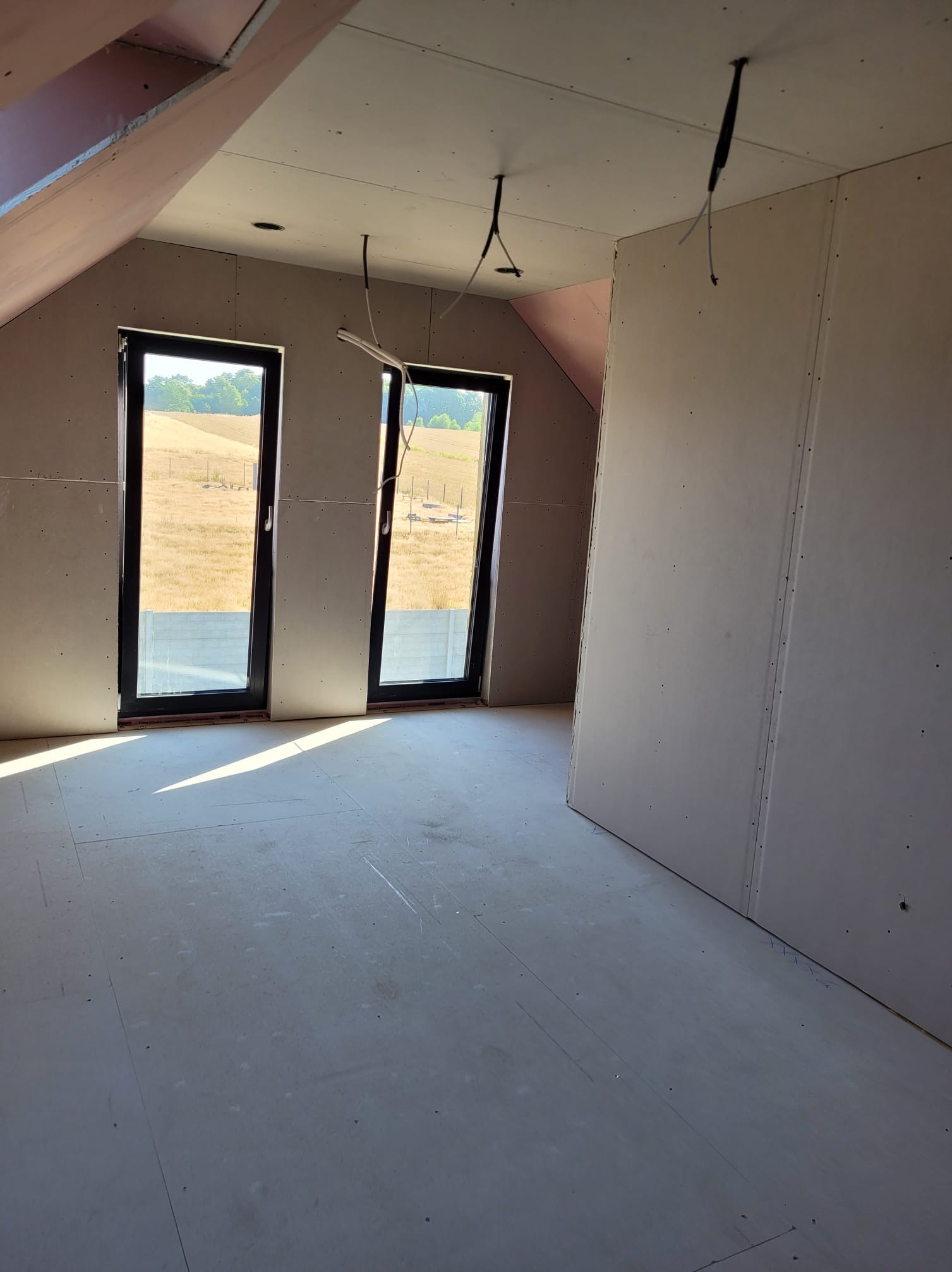 Poddasze w trakcie wykańczania: ściany obłożone płytami gipsowo-kartonowymi, dwa okna z widokiem na pole, wiszące kable elektryczne. Etap prac wykończeniowych.