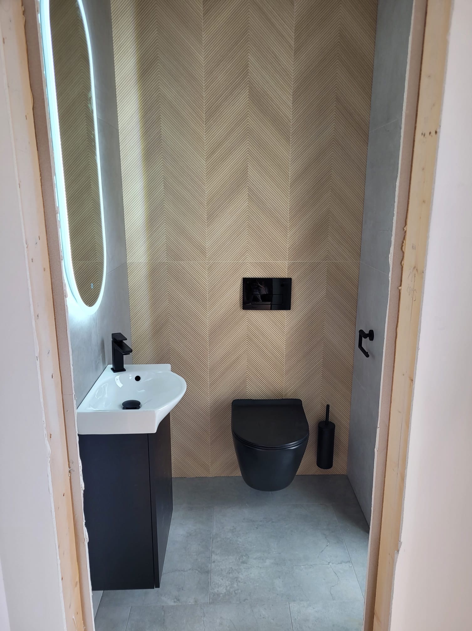 Nowoczesna łazienka z umywalką na szafce, toaletą w kolorze czarnym i lustrem z podświetleniem LED. Na ścianie płytki ułożone w jodełkę.