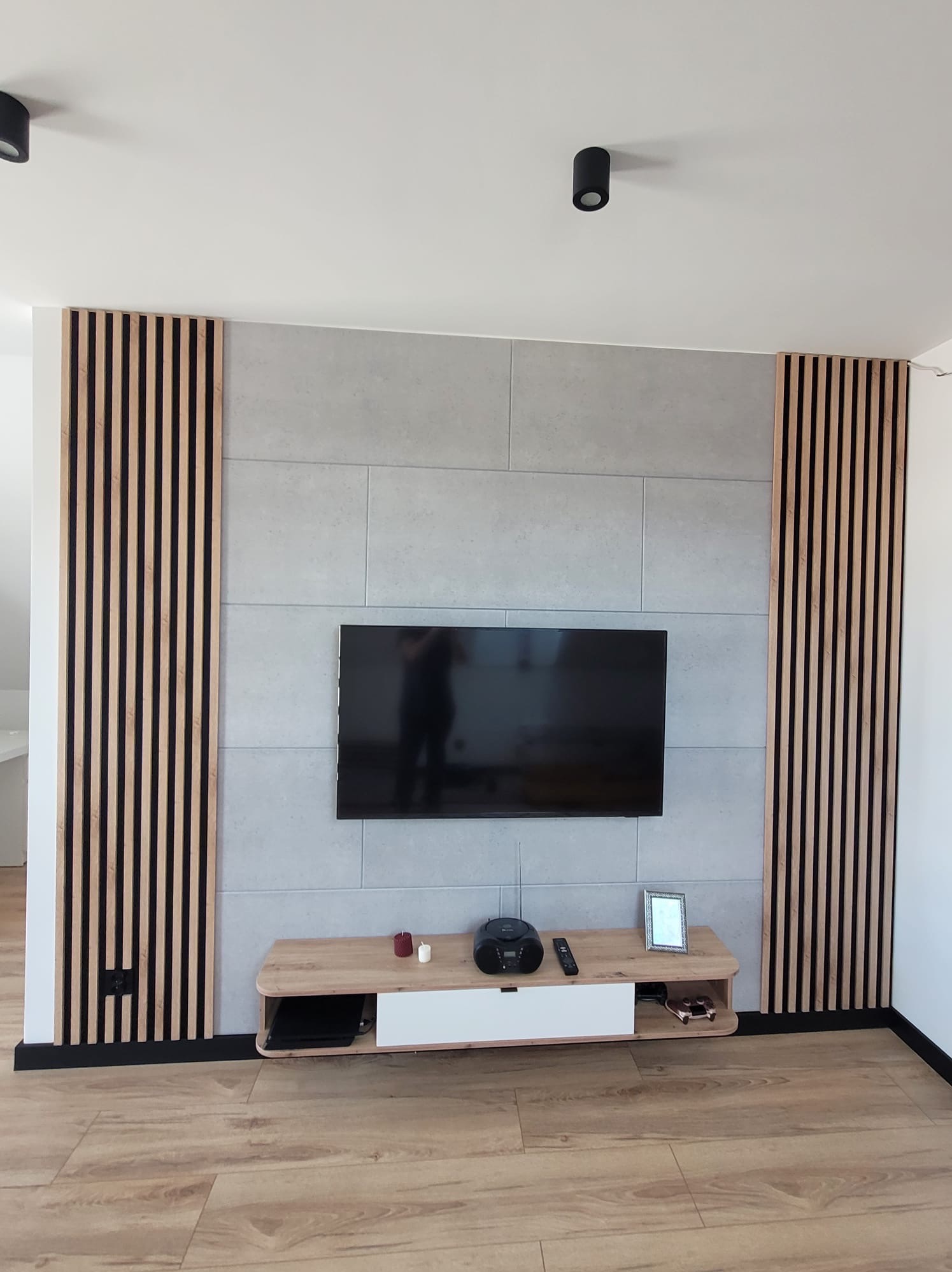 Nowoczesny salon z szarą ścianą z płytek, telewizorem, drewnianymi lamelami po bokach i szafką RTV. Podłoga z paneli imitujących drewno. Minimalistyczne oświetlenie.