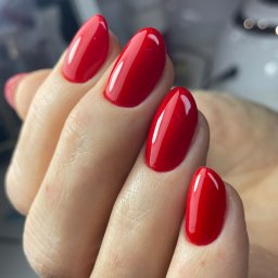 Saska Nails - Dłoń z czerwonym, błyszczącym lakierem hybrydowym na owalnych paznokciach. Widoczne delikatne refleksy światła na powierzchni lakieru, podkreślające jego gładkość.