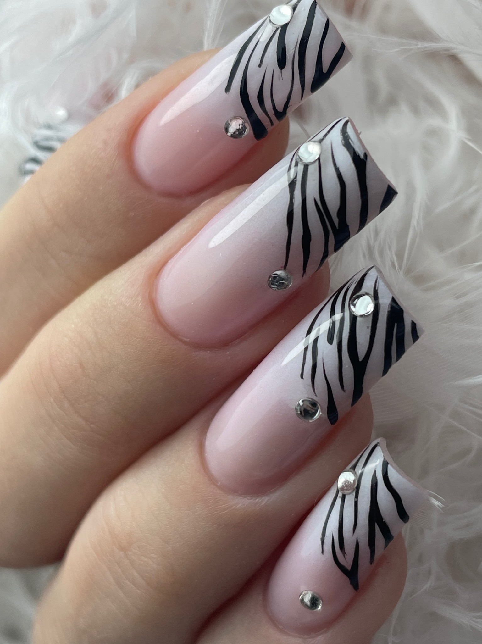 Dłoń z kwadratowymi paznokciami w odcieniach różu, zdobionymi wzorem zebry i cyrkoniami, na tle białych piór. Efektowny i modny design.