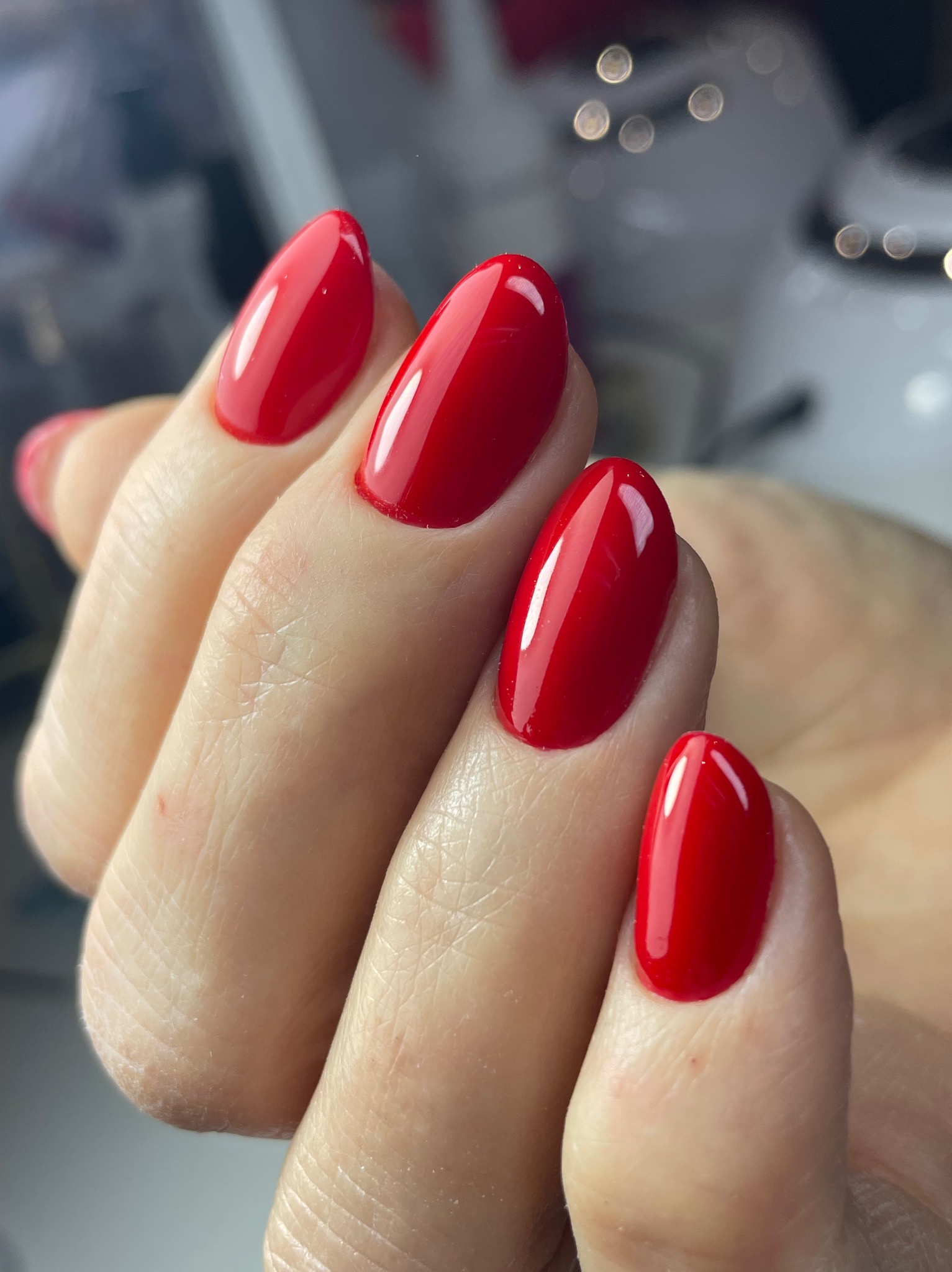Dłoń z czerwonym, błyszczącym lakierem hybrydowym na owalnych paznokciach. Widoczne delikatne refleksy światła na powierzchni lakieru, podkreślające jego gładkość.