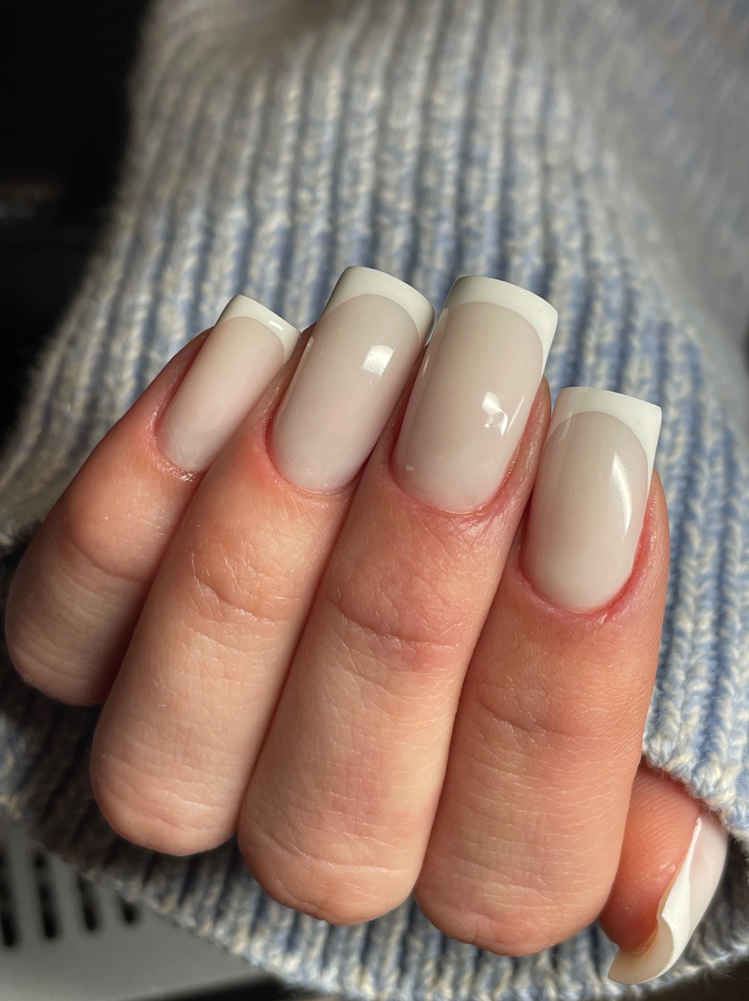 Dłoń z kwadratowymi paznokciami w stylu french manicure, na tle niebieskiego swetra. Delikatny odcień beżu i śnieżnobiałe końcówki. Precyzyjne wykonanie.