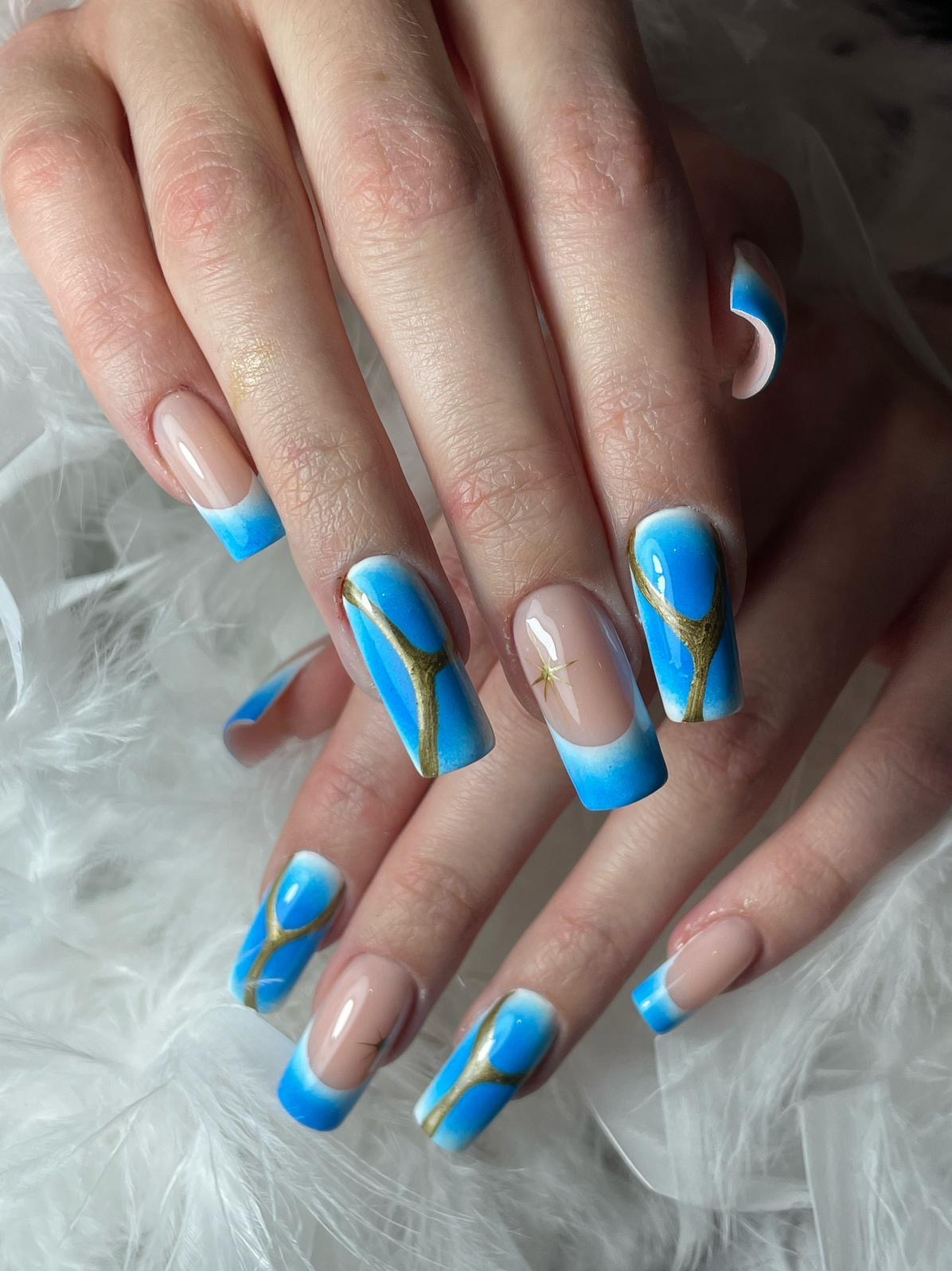 Dłonie z niebiesko-złotym manicure hybrydowym: ombre z geometrycznym wzorem na długich paznokciach, na białym, puszystym tle. Detal zdobienia.