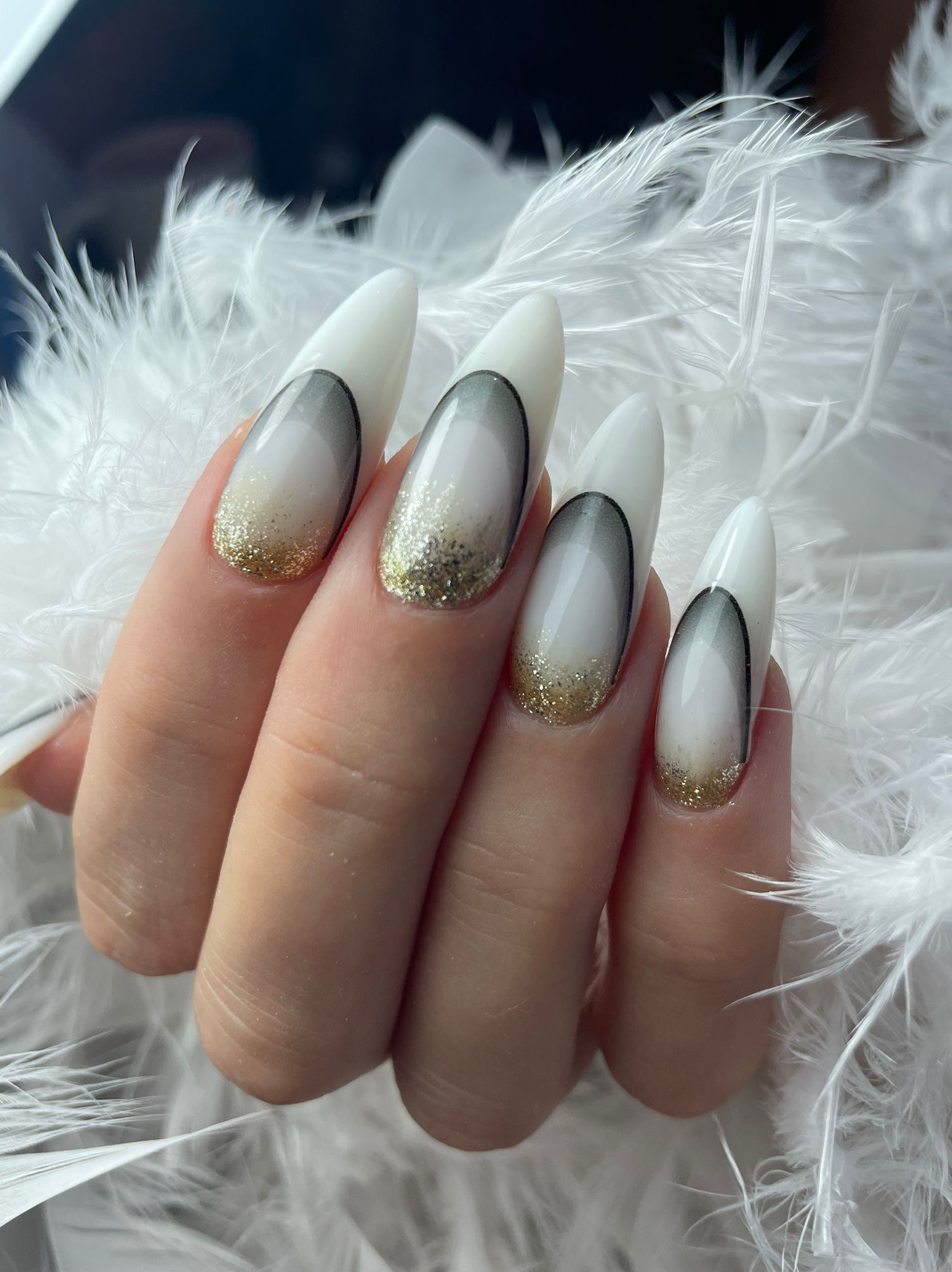 Dłoń z białymi, szpiczastymi paznokciami. Zdobienia: czarny łuk, biały gradient i złoty brokat u nasady. Tło z białych piór dodaje lekkości i elegancji.