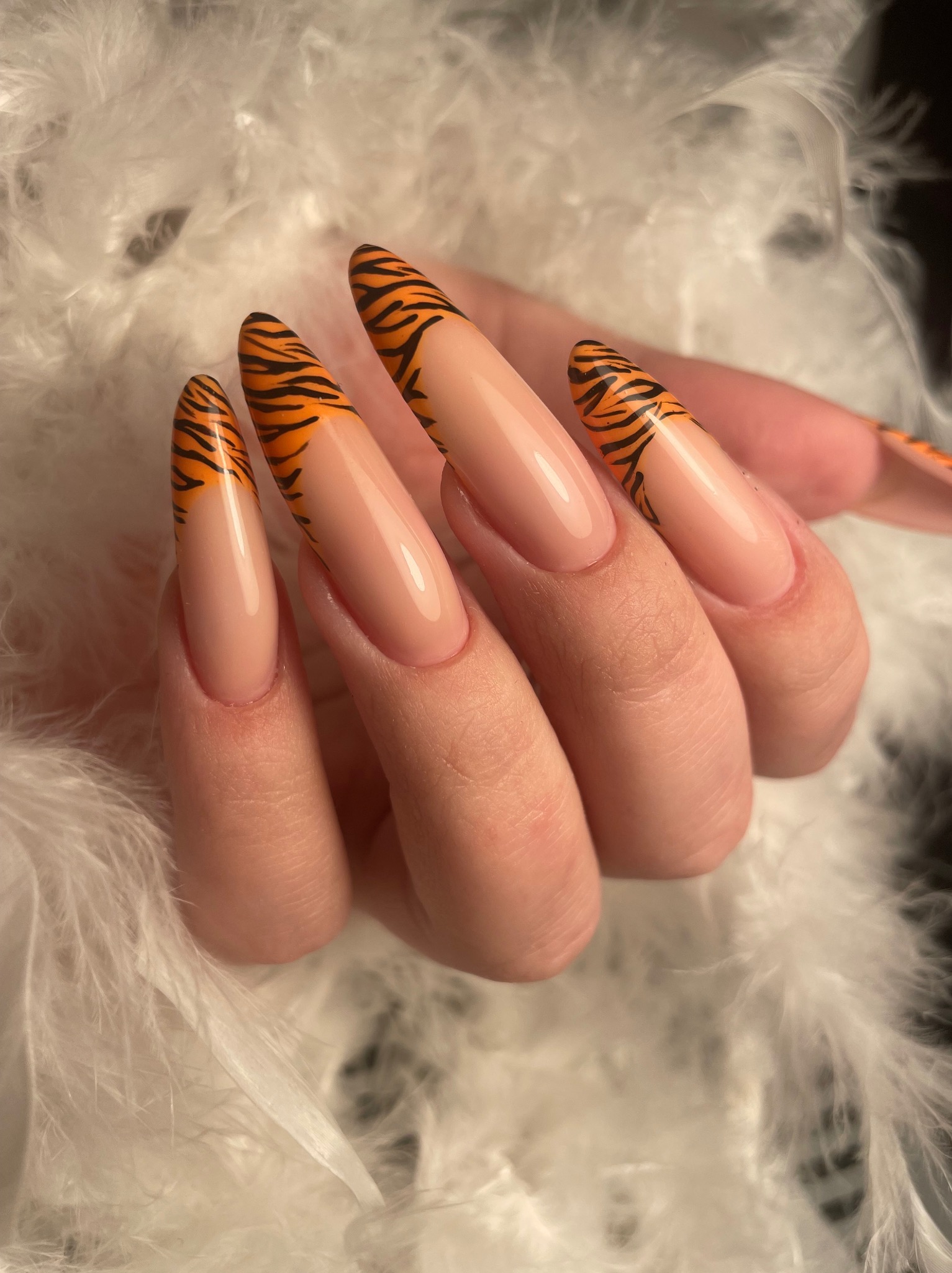 Dłoń z długimi, beżowymi paznokciami w kształcie migdała, ozdobionymi pomarańczowo-czarnym wzorem tygrysim na końcach, na tle białych piór.