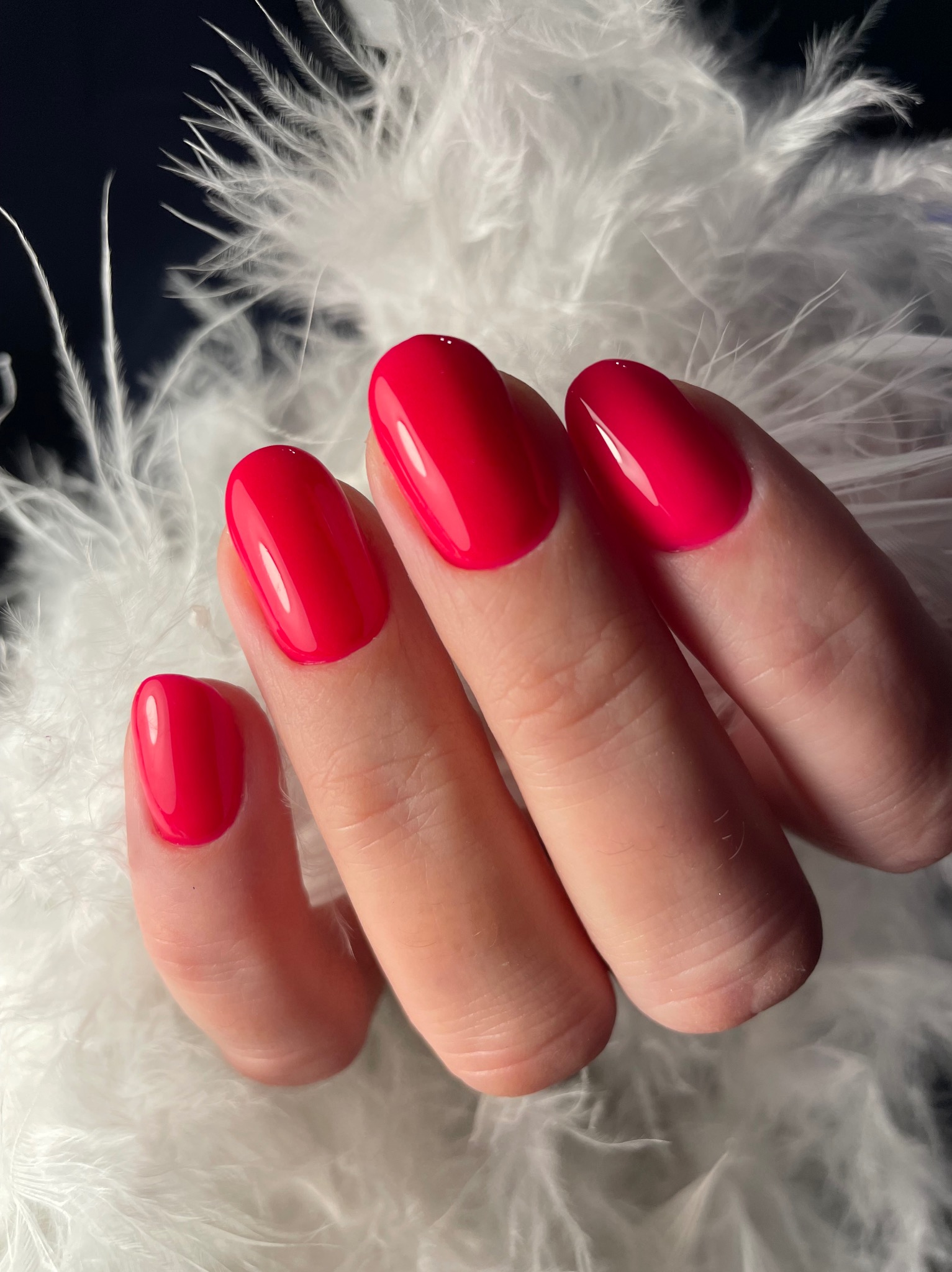 Dłoń z czerwonym, błyszczącym manicure hybrydowym na owalnych paznokciach, na tle białych, puszystych piór. Precyzyjne wykonanie, gładka powierzchnia lakieru.