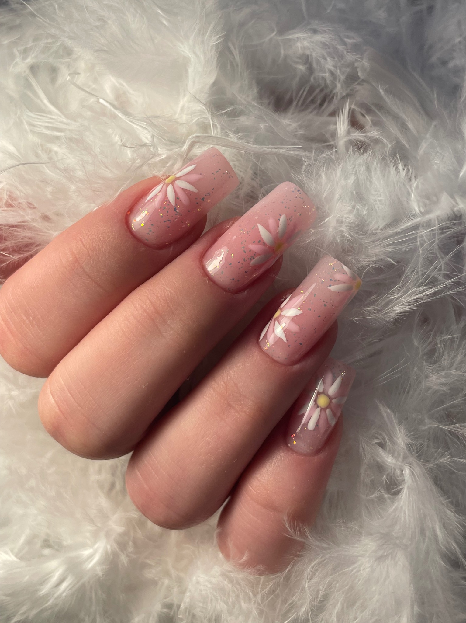 Ręka z kwadratowymi paznokciami w odcieniach różu, zdobionymi motywem białych kwiatów i złotym brokatem, na tle białych piór. Delikatny, dziewczęcy design.