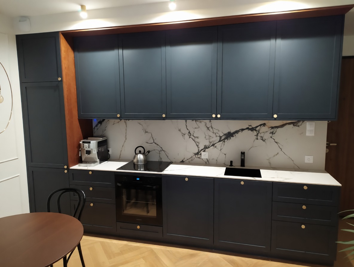 Granatowa zabudowa kuchenna z białym blatem i marmurowym panelem, złote gałki, widok na stół i krzesło, parkiet w jodełkę. Ekspres do kawy na blacie.