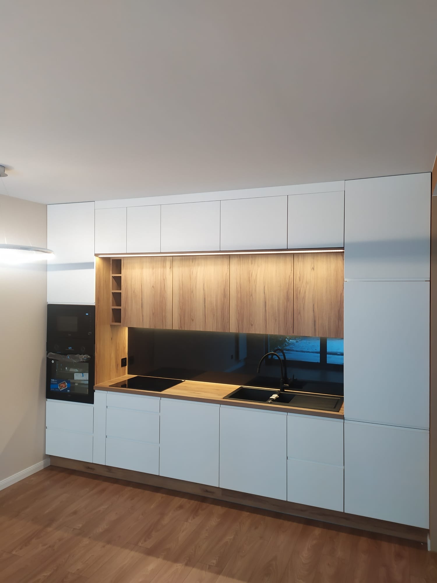 Nowoczesna zabudowa kuchenna w Łubiance: białe fronty, drewniane akcenty, czarny panel szklany i armatura. Minimalistyczny design, oświetlenie LED.