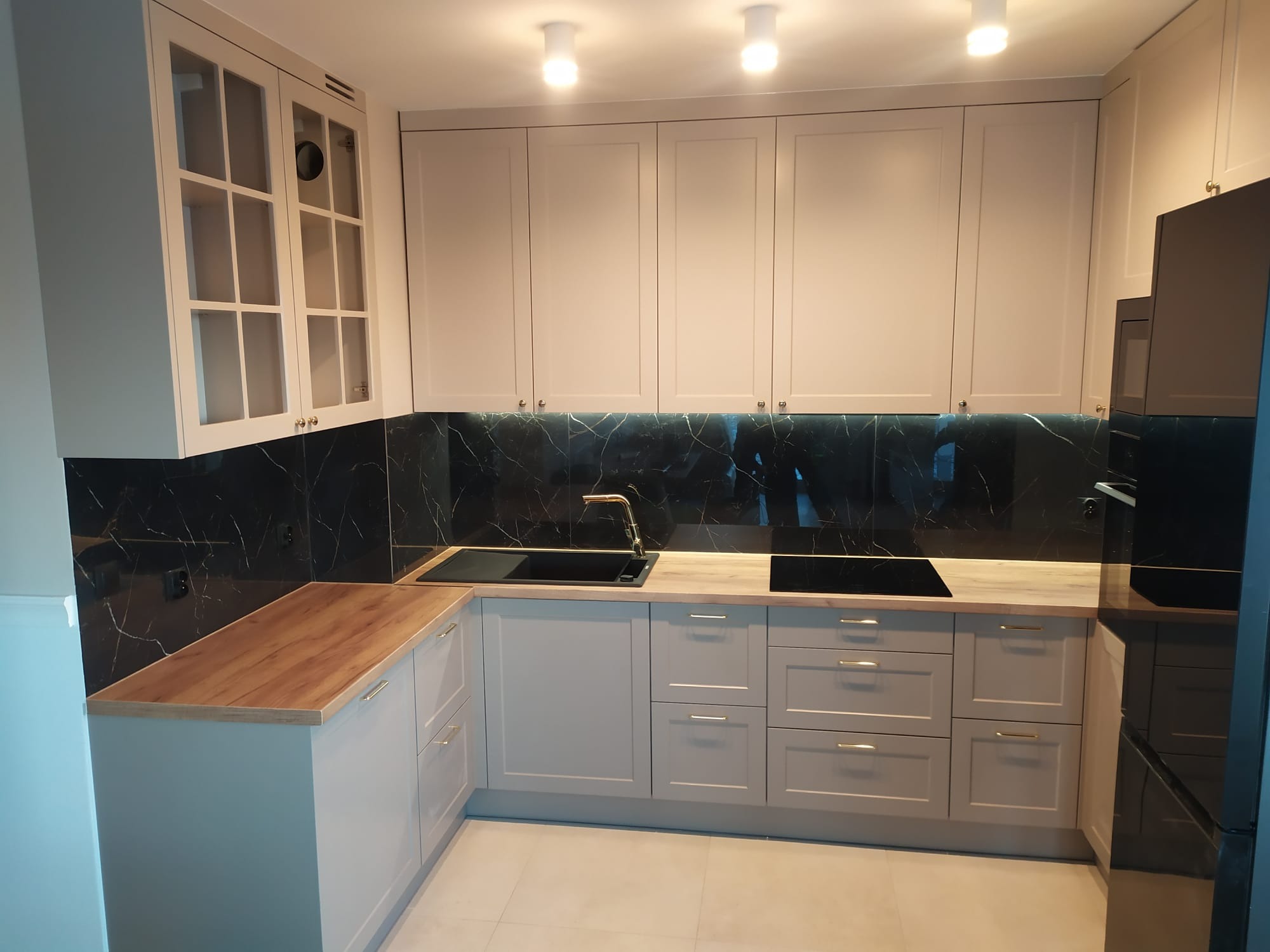 Jasna kuchnia z drewnianym blatem i czarnym marmurem na ścianie. Górne szafki z przeszkleniami. Złote uchwyty dodają elegancji. Nowoczesny design.