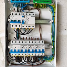 Rafał Barwikowski - Rozdzielnica elektryczna Schrack z zainstalowanymi bezpiecznikami i okablowaniem, widok z góry. Instalacja w białej obudowie na tle surowej ściany.