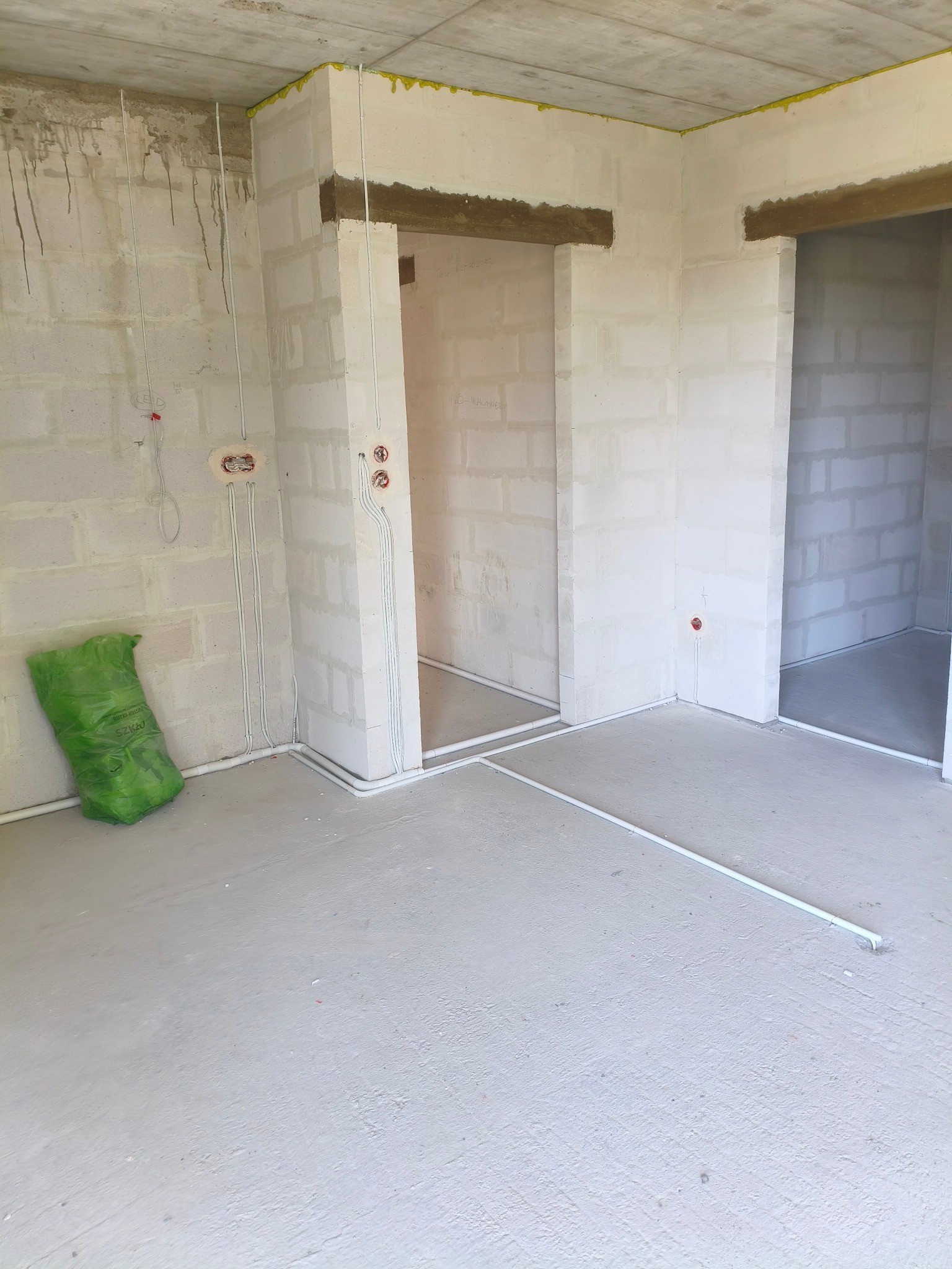 Surowy stan wnętrza z widocznymi przewodami elektrycznymi w białych rurkach na ścianach z betonu komórkowego i podłodze. Widoczne otwory drzwiowe.