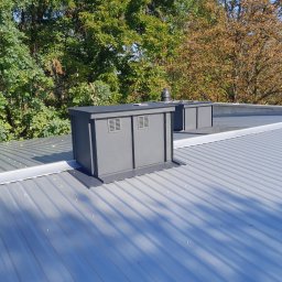 PHU Dachy - Paweł Wrzodak - Szara blacha trapezowa na dachu z dwoma kominami wentylacyjnymi. Widoczne mocowania blachy. W tle drzewa z zielonymi i brązowymi liśćmi.