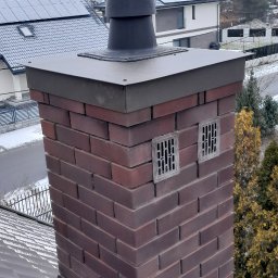Dachy Paweł Wrzodak - Komin z ciemnoczerwonej cegły z czarną nasadą wentylacyjną i dwoma kratkami, osadzony na dachu z widocznymi metalowymi uszczelnieniami. W tle domy z panelami fotowoltaicznymi.