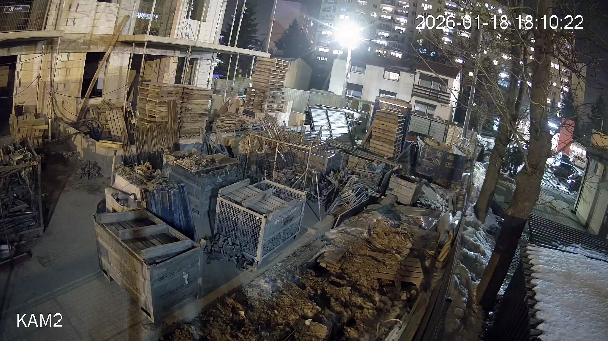 Nocny widok z kamery monitoringu na plac budowy z materiałami i konstrukcją w stanie surowym, oświetlony sztucznym światłem. Widoczne budynki w tle.