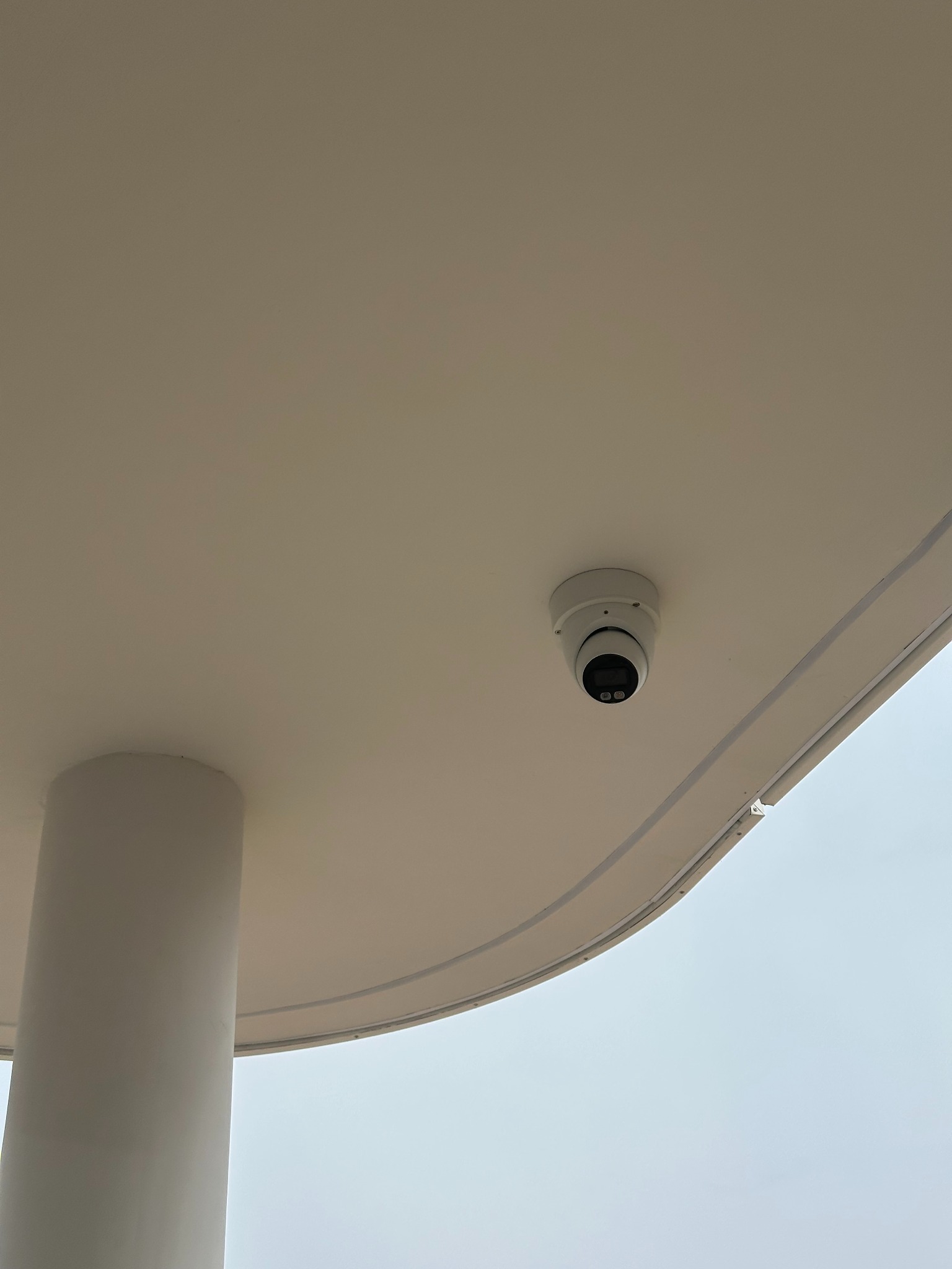 Kamera kopułkowa monitoringu zamontowana na suficie pod zadaszeniem, widok z dołu. Widoczny fragment kolumny i niebo w tle.