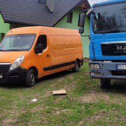 Mateusz Bujok - Pomarańczowy bus i niebieska ciężarówka MAN na trawie przed zielonym domem z ciemnym dachem. Widoczne tablice rejestracyjne pojazdów. Pochmurne niebo w tle.