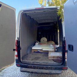 Mateusz Bujok - Otwarty tył białego busa z załadunkiem: panele, płytki i kartony. Widoczne otwarte drzwi, podłoga przestrzeni ładunkowej i fragment otoczenia zewnętrznego.