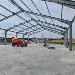SpecMontSerwis Radosław Suchecki - Konstrukcja stalowa hali w trakcie budowy, widoczny podnośnik koszowy i narzędzia na betonowej posadzce. Słupy i kratownica tworzą szkielet budynku.