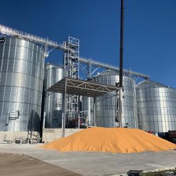 SpecMontSerwis Radosław Suchecki - Kompleks silosów zbożowych ze stali nierdzewnej w słoneczny dzień, z górą ziarna u podstawy. Widoczna konstrukcja wsporcza i ciężarówka w tle.