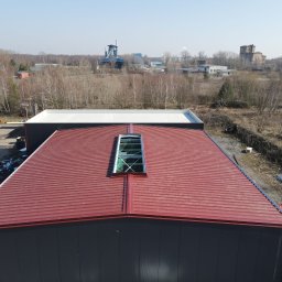 SpecMontSerwis Radosław Suchecki - Hala z bordową blachą trapezową i świetlikiem dachowym, widok z góry. W tle zabudowania przemysłowe i las. Na placu zaparkowane autobusy i podnośnik koszowy.